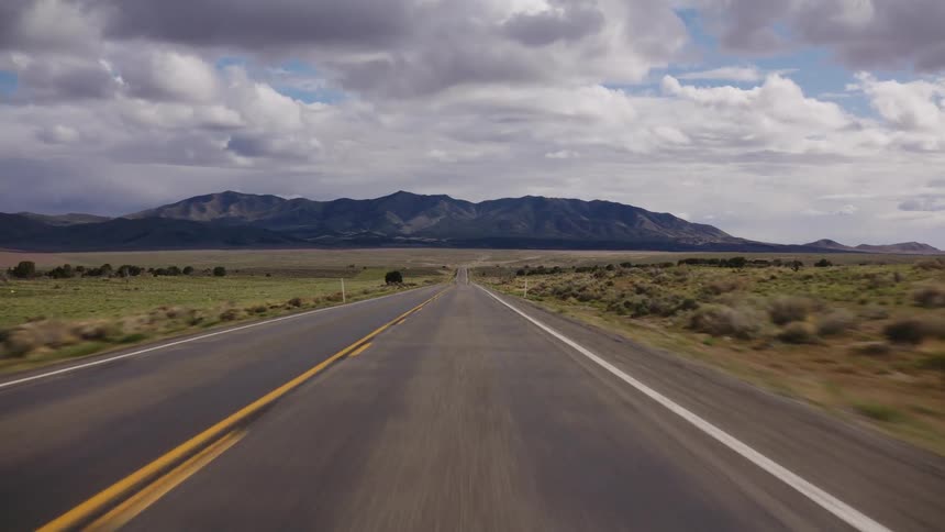 A point-of-view shot of driving down a long, straight desert highway towards a distant mountain range under a cloudy sky