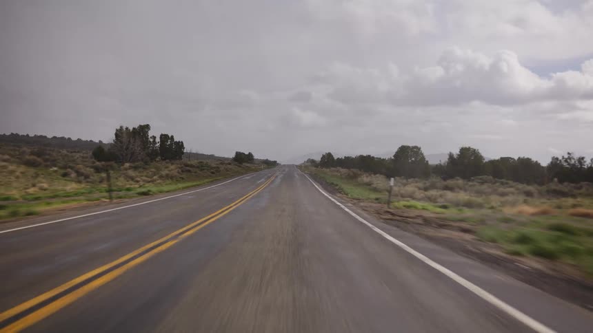 A driver's point-of-view traveling down a wet and empty two-lane highway through the countryside on a cloudy day