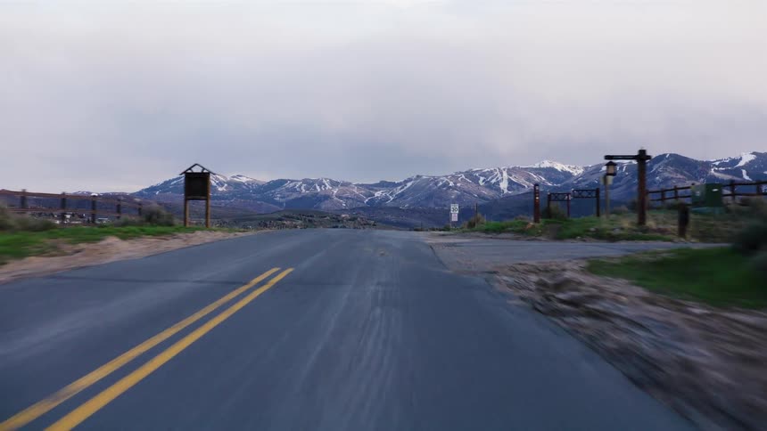 Driving along a quiet country road toward a majestic snow-capped mountain range under a cloudy sky