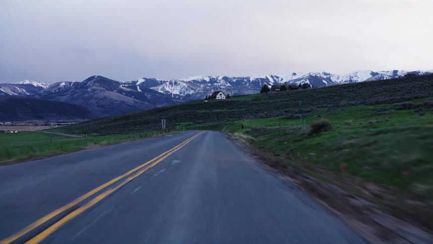 A first-person perspective driving along a remote country road towards snow-capped mountains at dusk