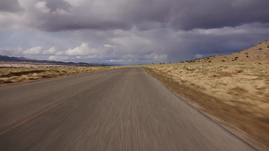 POV driving fast down a remote road through an arid landscape under dramatic storm clouds