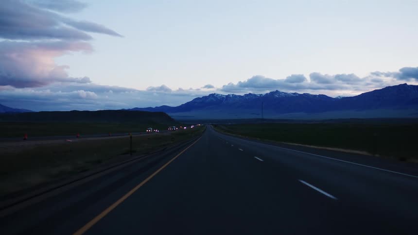 A point-of-view shot captures a serene drive along a highway at dusk, with traffic in the distance and snow-capped mountains on the horizon