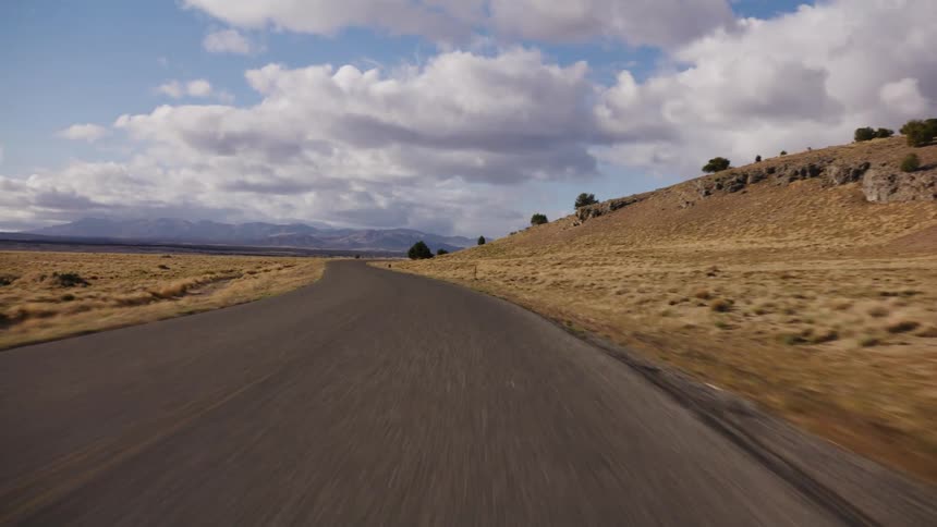 A point-of-view drive on a winding road through an arid, hilly landscape under a partly cloudy sky