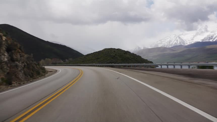 Driving POV on a scenic mountain highway next to a lake with snowy peaks under a cloudy sky