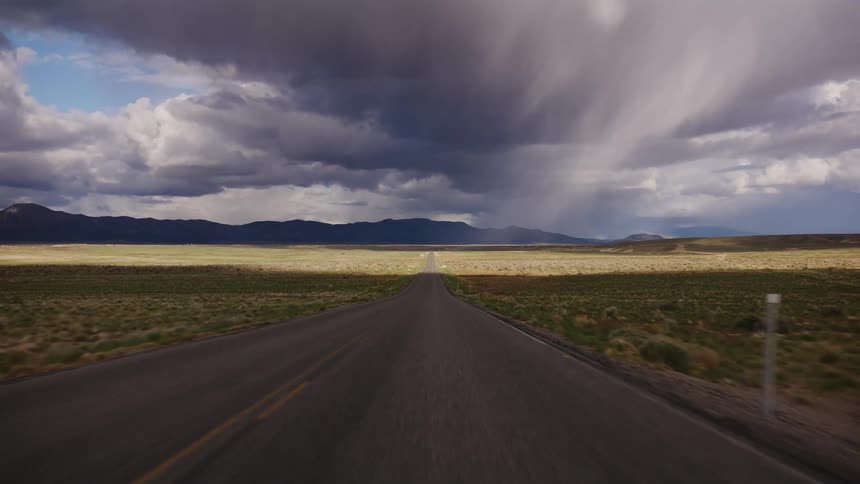 A POV shot moving down a remote highway through a vast valley with a powerful storm brewing ahead