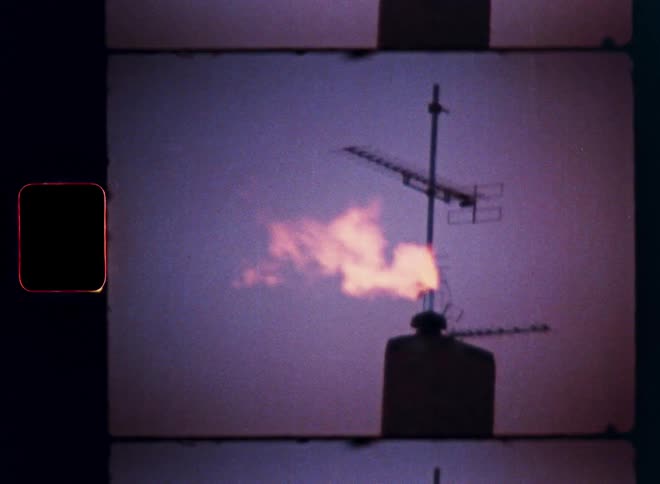 A vintage Super 8 film captures a gas flare burning from a rooftop pipe next to an antenna against a purple dusk sky