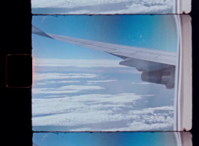 A vintage film shot from an airplane window shows the wing flying high above a sea of clouds
