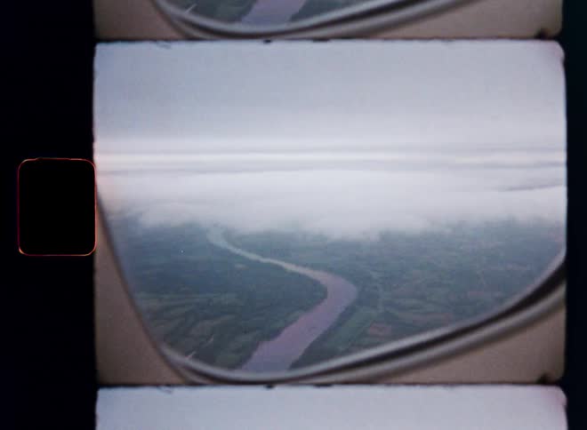 Vintage film footage from an airplane window captures an aerial view of a winding river through a green landscape below the clouds