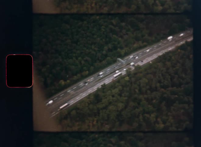 Vintage 8mm footage captures an aerial view of cars and trucks traveling on a highway surrounded by woods