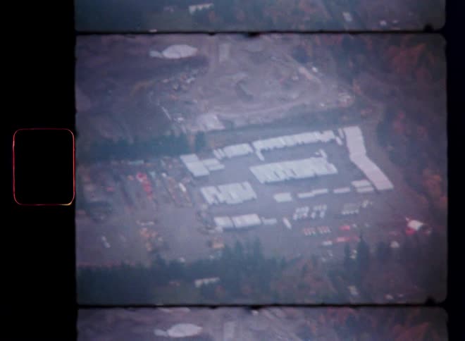An old, grainy aerial shot on Super 8 film reveals a large industrial yard surrounded by trees