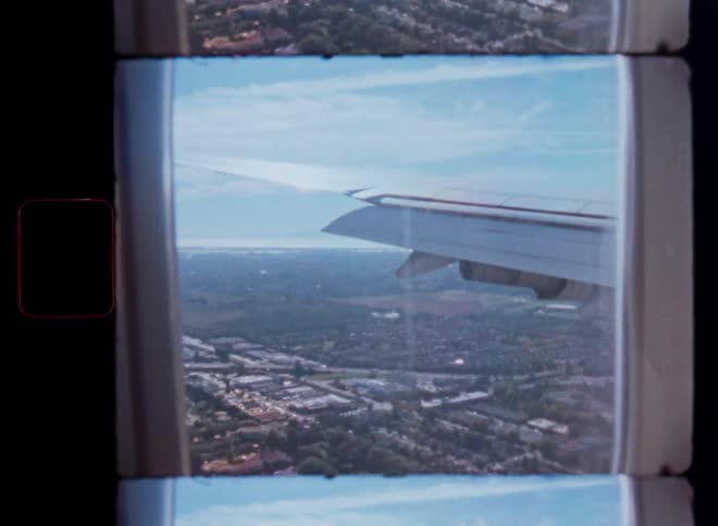 A vintage film view from an airplane window shows the wing flying over a sprawling city towards the distant coast