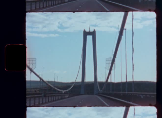 A vintage film shot captures the view from a car driving over a massive suspension bridge on a sunny day