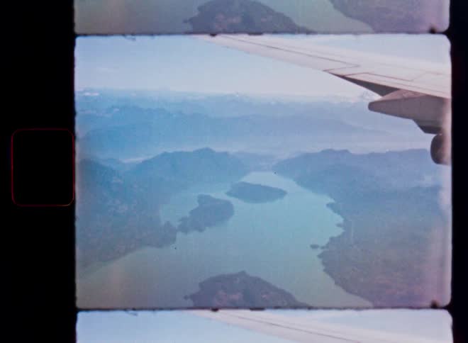 An airplane window view reveals a vast, hazy mountain landscape with a winding river below, captured on vintage 8mm film