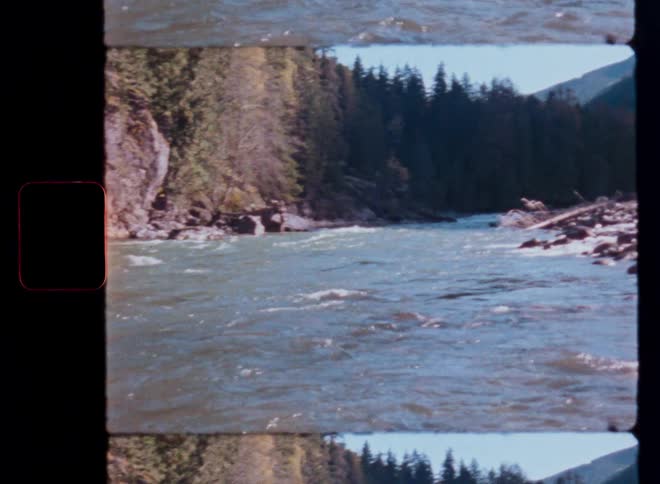 Vintage film footage pans across a fast-flowing river cutting through a rocky, forested mountain valley on a sunny day