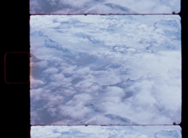 An aerial view from an airplane window captures a majestic, snow-covered mountain range peeking through the clouds on vintage film