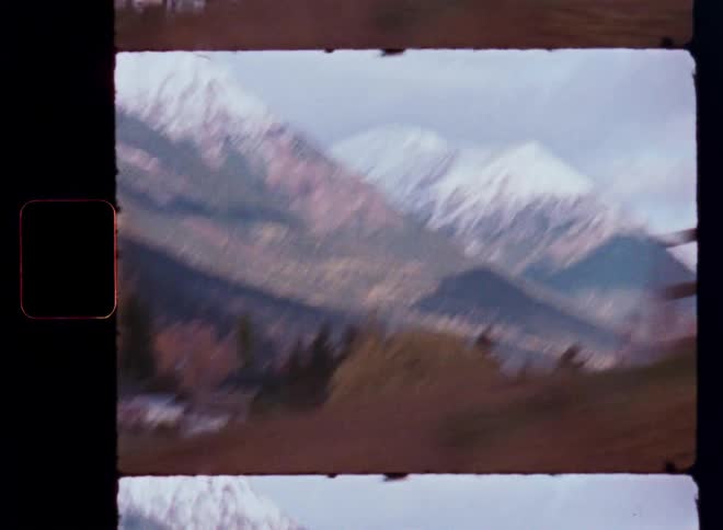 Vintage 8mm film captures a scenic drive past majestic snow-capped mountains and an autumnal forest village