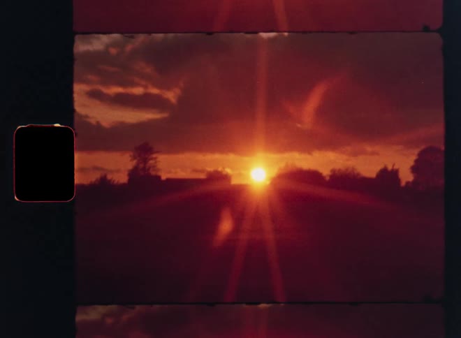 A nostalgic Super 8 film captures a brilliant red sunset over a silhouetted treeline on the horizon