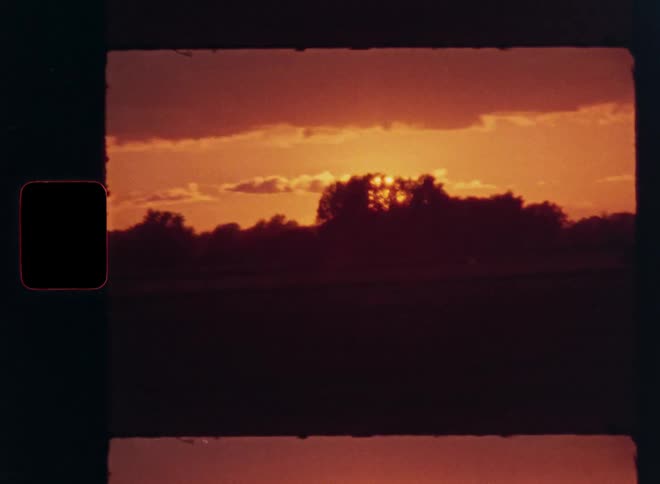 Vintage film captures a dramatic time-lapse of the sun setting behind dark clouds and a silhouetted landscape under a fiery red sky