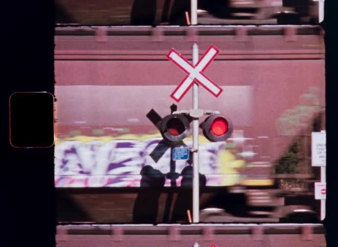 A close up of a flashing red railroad crossing light as a long freight train with graffiti speeds by, shot on vintage film