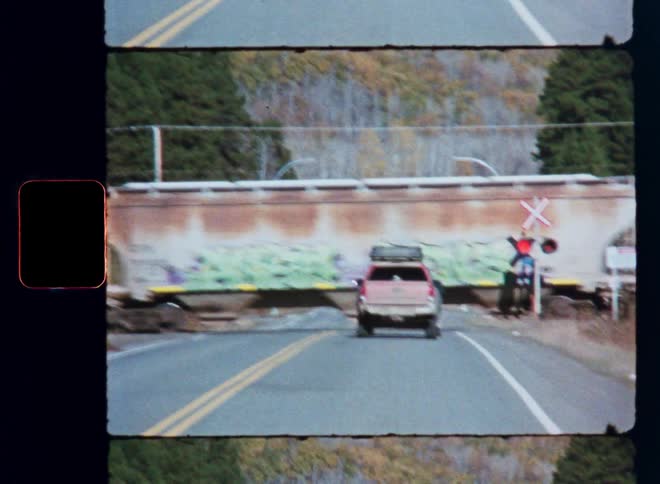 A pickup truck waits at a rural railroad crossing as a long freight train covered in graffiti slowly passes by