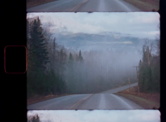 A vintage Super 8 film captures a moody shot of a road leading into a foggy forest with mountains in the background
