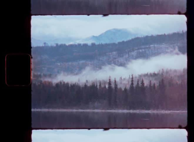 A serene Super 8 film shot of fog moving through a snowy mountain forest reflected in a still lake