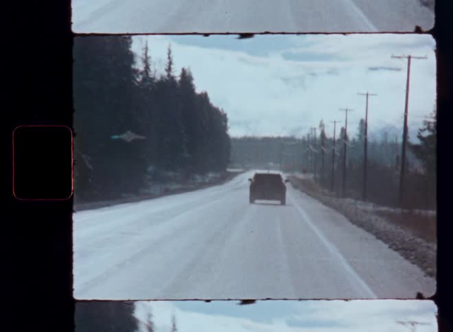 A vintage point-of-view shot follows a car driving down a snowy highway through a winter landscape with mountains in the distance