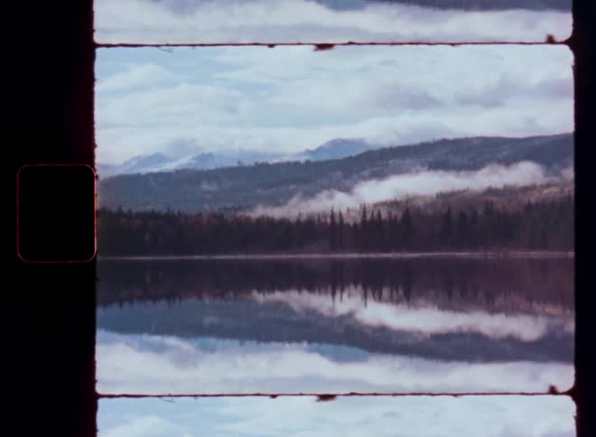 Vintage 16mm film scan of a calm lake perfectly reflecting the foggy mountains and cloudy sky