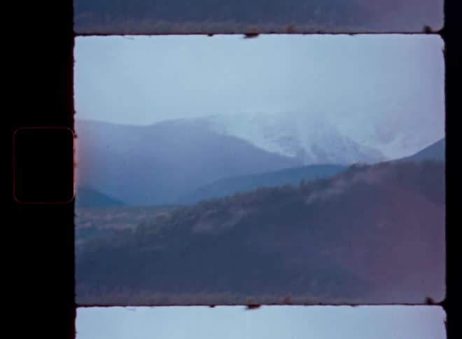 A vintage film frame captures a serene, overcast day with snow-capped mountains looming over a dense, hilly forest