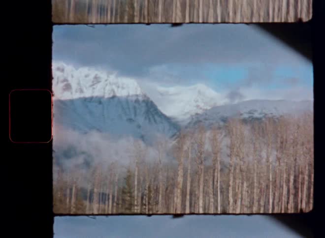 A vintage film gate scan shows a moving shot of a snow-capped mountain range with a foggy valley and a forest of trees in the foreground