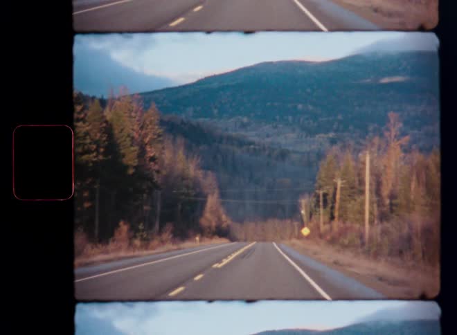 An authentic vintage film shot captures the view from a car driving down a quiet rural road towards distant mountains