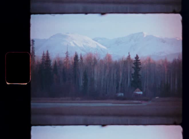 Vintage super 8 footage shows a tranquil Alaskan landscape with a forest and lake at the base of snowy mountains, ending with a vibrant film burn