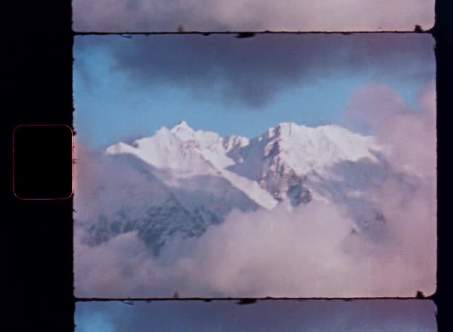 A vintage 16mm film shot captures a majestic snowy mountain peak emerging through a layer of soft, drifting clouds