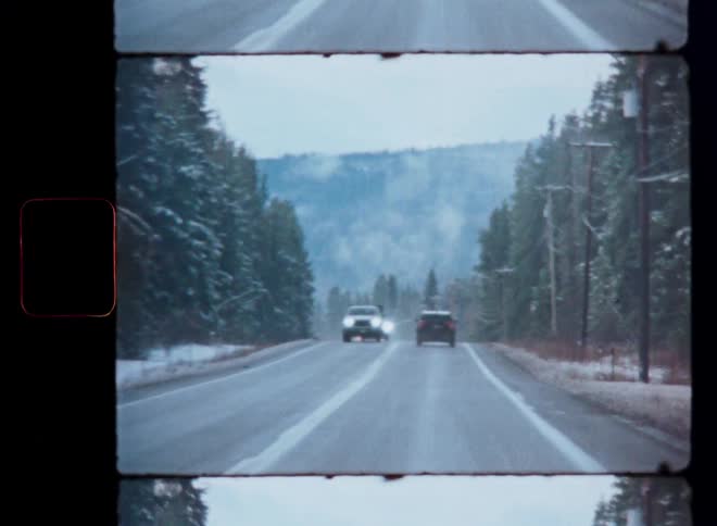 A retro-style, point-of-view shot captures cars driving down a two-lane road through a snowy evergreen forest in the mountains