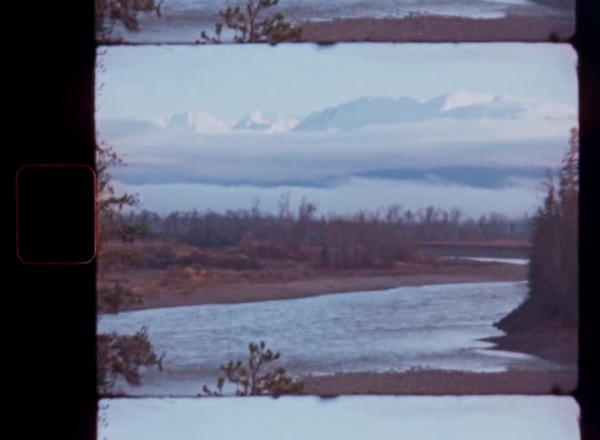 Vintage film footage captures a serene river flowing with snow-capped mountains rising above the clouds in the distance