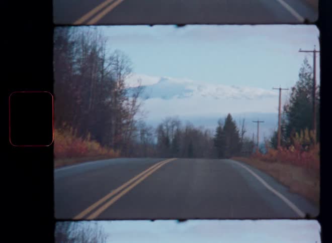 A nostalgic handheld film shot captures a quiet rural road leading towards a distant snow-capped mountain