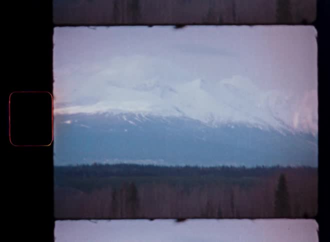 A slow pan reveals a majestic, snow-covered mountain range towering over a vast forest, captured on vintage 8mm film