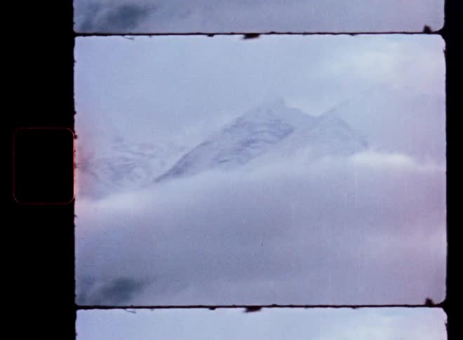 A vintage film captures the majestic, snow-capped peaks of a mountain range emerging from thick, rolling clouds