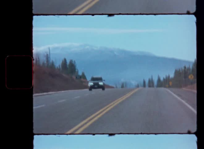 A vintage 8mm film shot of a black SUV driving on a mountain highway with snowy peaks in the background