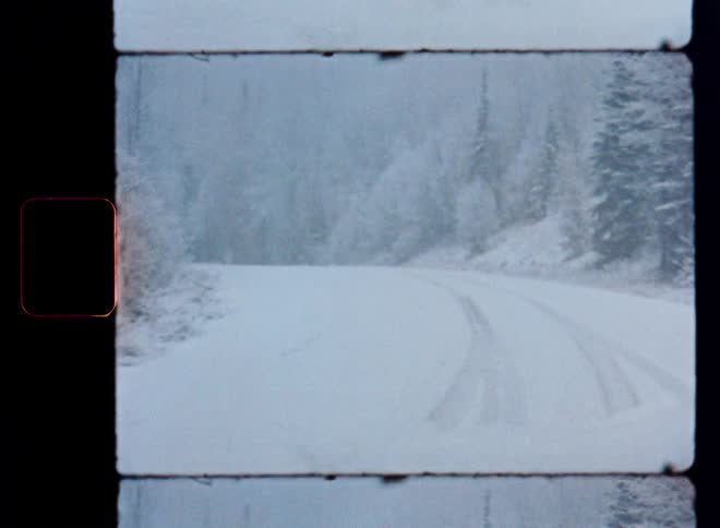 A slow point-of-view drive down a winding, snow-covered road through a forest during a gentle snowstorm, captured on vintage film