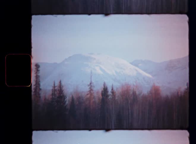 Vintage 8mm film reveals a tranquil, snow-covered mountain rising above a quiet forest landscape