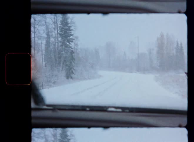 A vintage home movie captures the view from a car driving slowly down a snow-covered road during a blizzard