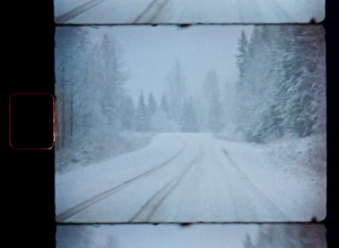 Vintage 8mm film captures a lonely, snow-covered road disappearing into a forest during a heavy snowstorm