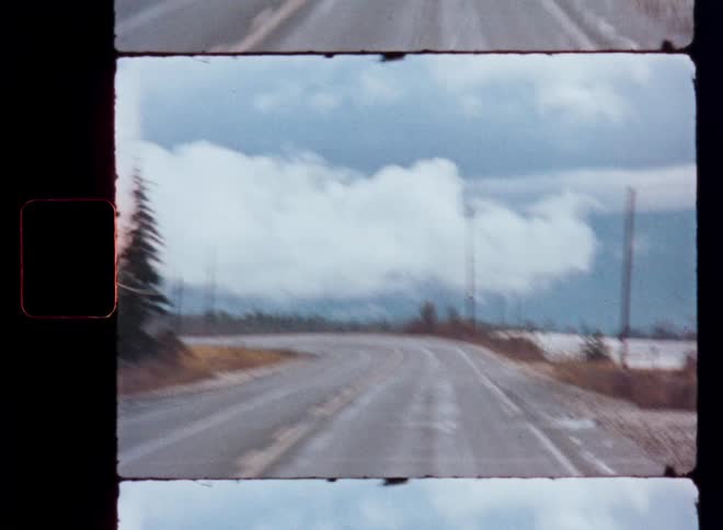 Vintage 8mm film captures the view from a car driving along a winding road on a cold, cloudy winter day