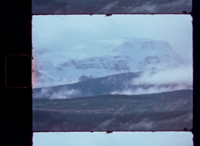 A vintage film scan shows a moody landscape of snow-capped mountains partially obscured by rolling fog and clouds