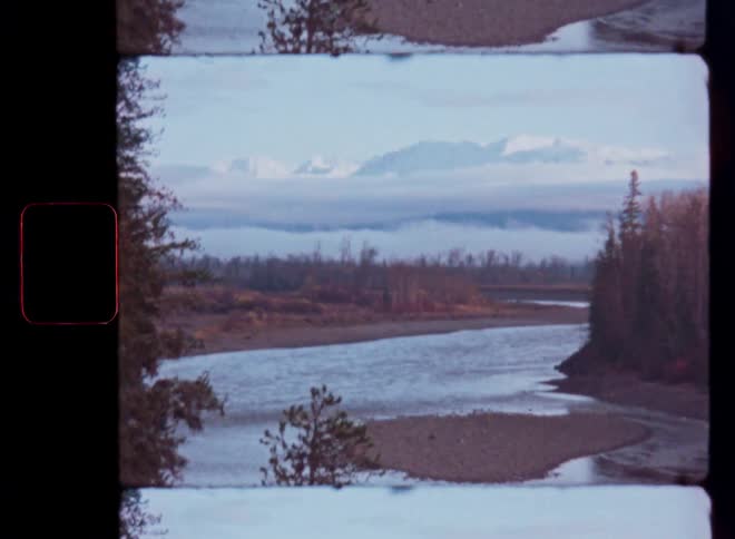 Super 8 film scan reveals a tranquil river surrounded by trees with majestic snowy mountains appearing through the clouds