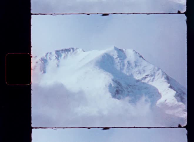 An upward tilt on vintage film reveals a stunning, snow-covered mountain peak emerging from the clouds