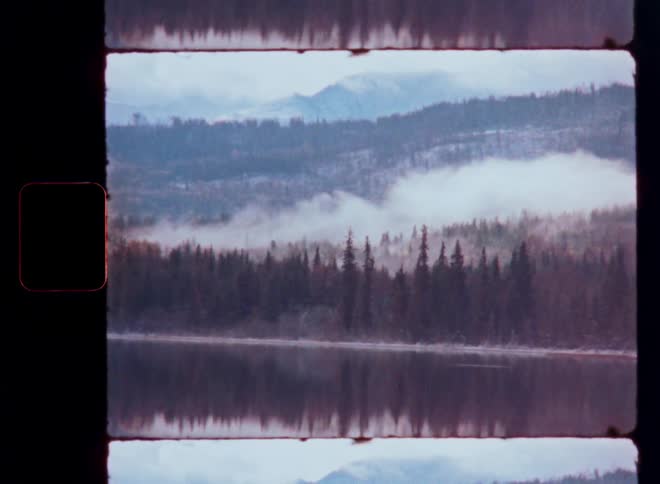 A moody and tranquil vintage film clip shows a still lake perfectly reflecting a foggy pine forest and distant mountains