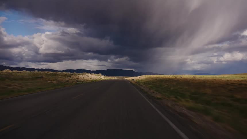 A first-person perspective driving along a desolate country road toward a dramatic storm cloud and rain falling in the distance