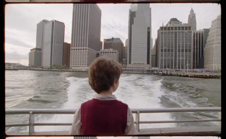 A young boy watches the New York City skyline disappear into the distance from the back of a moving ferry in this vintage-style shot